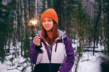 A young woman is celebrating Christmas in a snowy forest, holding a sparking sparkler.