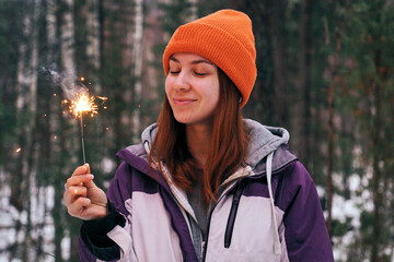 A young woman is celebrating Christmas in a snowy forest, holding a sparking sparkler.