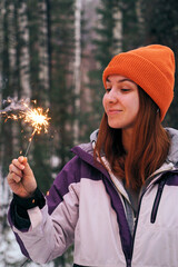 A young woman is celebrating Christmas in a snowy forest, holding a sparking sparkler.