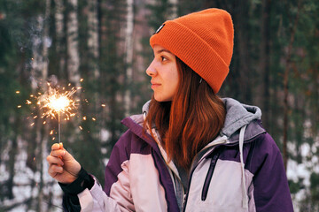 A young woman is celebrating Christmas in a snowy forest, holding a sparking sparkler.
