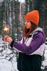 A young woman is celebrating Christmas in a snowy forest, holding a sparking sparkler.