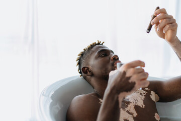 Young african american man with vitiligo looking at cigar and holding glass while taking bath at...