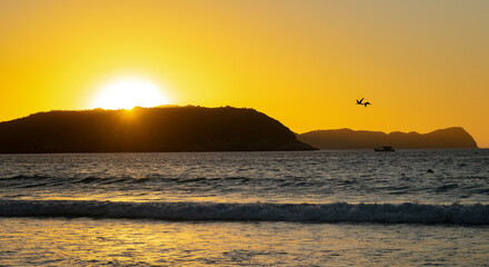 Sunrise at Praia do Forte, boat, the sun coming out from behind the mountains and illuminating the...