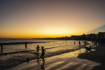 Sunset, with the last rays of sunlight leaving a beautiful orange hue in the sky behind the city of Cabo Frio and in the sea waters full of swimmers. Rio de Janeiro, Brazil-Editar