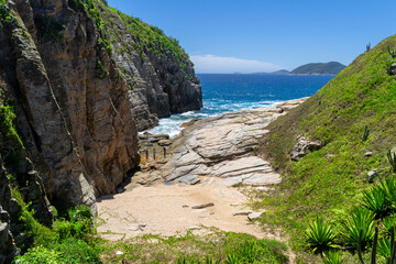The back of Praia das Conchas, with rock walls and waves crashing over them, vegetation, blue sky and islands in the distance. Cabo Frio City, Rio de Janeiro.