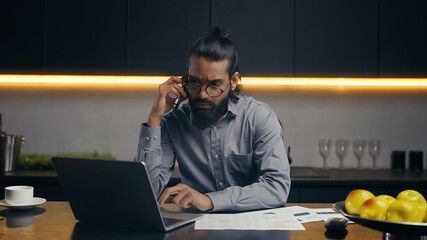 Young Indian businessman talking on phone, reviewing reports, typing on laptop - Powered by Adobe