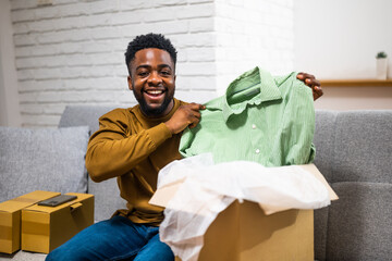 Happy African American man unpacking delivered package with new clothes while sitting on sofa at his cozy home.	