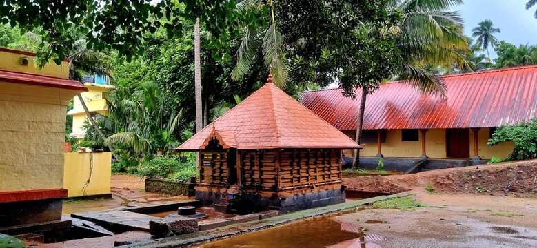 Intricate stone pillars and a vibrant red roof characterize the devotional atmosphere of Aryankavu Ayyappan Temple, capturing its unique architectural charm and rainy season tranquility amidst green