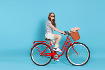 Beautiful teenage girl in sunglasses riding bicycle with basket of flowers against light blue...