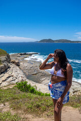 Woman at the viewpoint of Praia das Conhas, lots of rocks and crystal clear waters, blue sky and mountains in the background. Rio de Janeiro, Brazil.