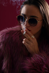 Beautiful woman in fur coat smoking cigarette on dark pink background, closeup