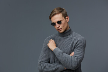 Stylish young man wearing gray turtleneck sweater and black sunglasses posing against plain gray...