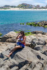 Woman sitting on a large rock, crystal clear sea, blue sky and city in the background. Praia Grande, Rio de Janeiro, Brazil.