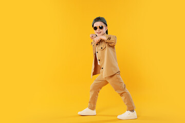 Happy little boy in sunglasses and baseball hat dancing on orange background. Space for text