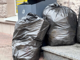 Piles of black trash bags are leaning against a stone surface next to recycling bins on a busy city...