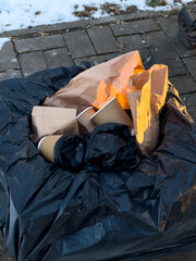 Discarded paper and coffee cups fill a black trash can near a snow-covered park area, highlighting...