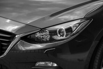 A close-up, high-angle black and white shot of a modern car's sleek headlight and front fender,...