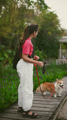 A young girl stands on a bridge in a park with her corgi dog and holds a bag of dog poop in her...