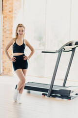 Fototapeta premium Active athletic woman in activewear standing confidently near a treadmill in a modern well-lit gym interior space