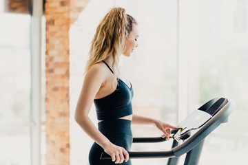 Athletic woman in activewear operates treadmill console at a fitness gym in bright natural lighting...
