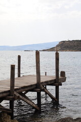 Old wooden pier over calm sea water with rocky shore in the foreground and mountains in the distance. Rustic coastal scene, peaceful and nostalgic atmosphere.
