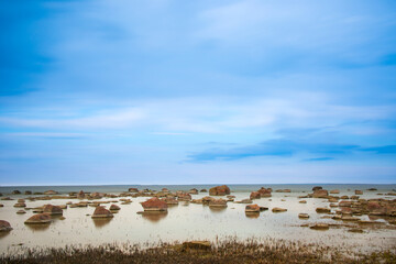 A serene shallow shoreline with scattered rocks, calm reflective water, and a soft blue sky...