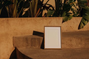 Minimalist elegant summer poster mockup in warm golden sunlight.. Empty vertical wooden picture frame on rustic concrete steps surrounded by tropical green plants. Palm, banana leaves. Branding design