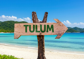 A wooden arrow sign points towards tulum on a tropical beach
