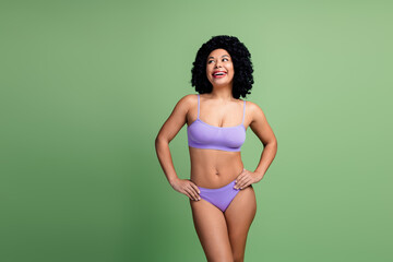 Confident young woman in purple lingerie posing with a smile against a green backdrop, celebrating...