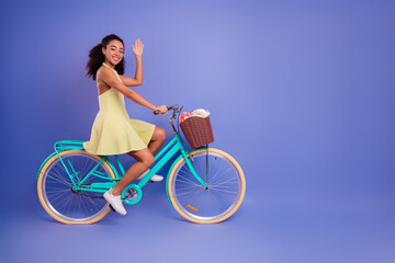 Young woman in yellow dress riding a blue bicycle with basket against plain purple backdrop waving...