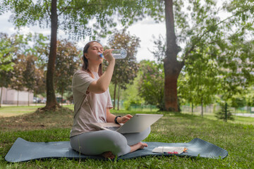 Female freelancer pausing from laptop work to hydrate seated cross-legged on yoga mat among grass...