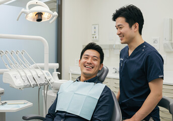 Obraz premium young asian patient in clinic, dentist wearing navy scrubs, man smiling in dental chair. modern equipment, relaxed atmosphere. healthcare, medical service, healthcare, stomatology.
