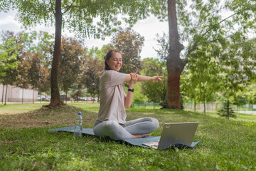 Relaxed young woman doing side arm stretch while seated on yoga mat with laptop, enjoying outdoor...