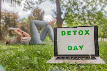 Clear text DETOX DAY on laptop screen green grass front woman lying back legs up reading book...