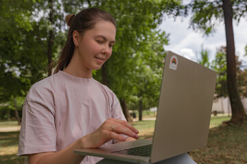 Woman seated using silver laptop green park background leafy trees daylight natural relaxed...