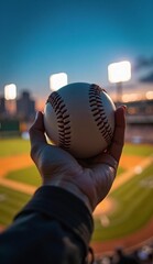 A hand holds a baseball against the backdrop of a brightly lit stadium at sunset.