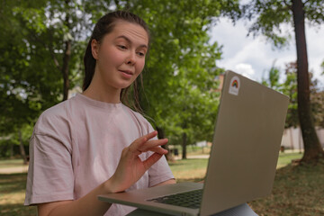 Woman with long hair in pale pink shirt gestures while sitting outdoors with laptop during video...