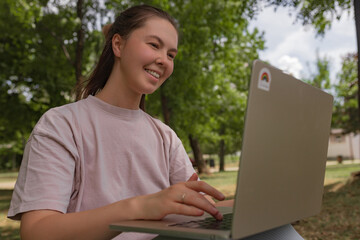 Woman in pink shirt smiling at laptop while typing outdoors, natural green park background.	Relaxed...