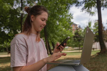 Woman with long hair in pale pink shirt multitasks outdoors using laptop while checking phone...