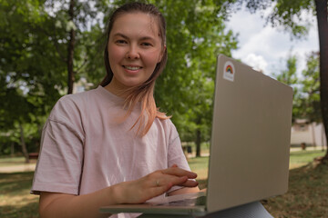 Woman in pink shirt with laptop outdoors smiling directly at camera, surrounded by green park...