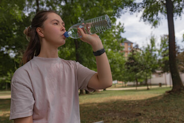 Woman in pale pink shirt with relaxed expression sips water from clear plastic bottle outdoors,...