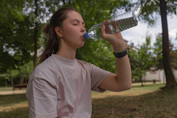 Woman in pale pink shirt closing eyes while drinking water from clear plastic bottle seated in...