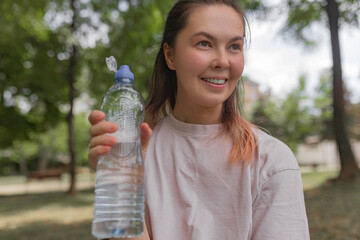 Woman holding clear plastic sport water bottle with open blue cap smiling while standing outdoors...