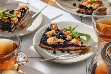 A slice of homemade blueberry crumble pie on a plate on white tile background. Sweet breakfast composition in bright morning light