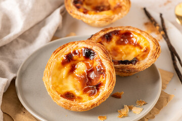 Three Pastel de Nata, Portuguese custard tarts, are presented on a plate with cinnamon and vanilla beans.