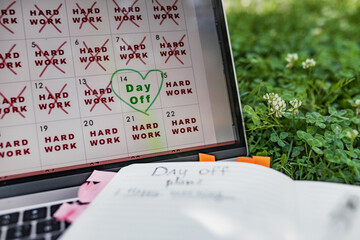 Close view of digital calendar filled with crossed 'HARD WORK' days, one green-marked break day stands out, positioned next clover flowers in sunlight, expressing nature work balance.