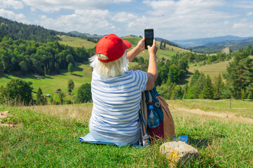 An active senior woman sits on a grassy hillside, taking a selfie with her smartphone while enjoying a peaceful view of rolling green hills and distant mountains. It's a sunny summer day, perfect for