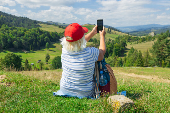 An active senior woman sits on a grassy hillside, taking a selfie with her smartphone while enjoying a peaceful view of rolling green hills and distant mountains. It's a sunny summer day, perfect for
