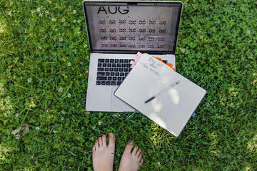 Relaxed barefoot setting with feet in soft grass, a laptop calendar, and a notebook open to 'Day...