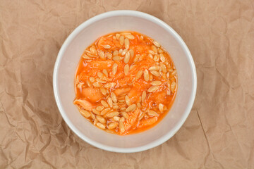 A white bowl is filled with a mixture of orange cantaloupe flesh and numerous seeds, set against a crumpled brown paper background